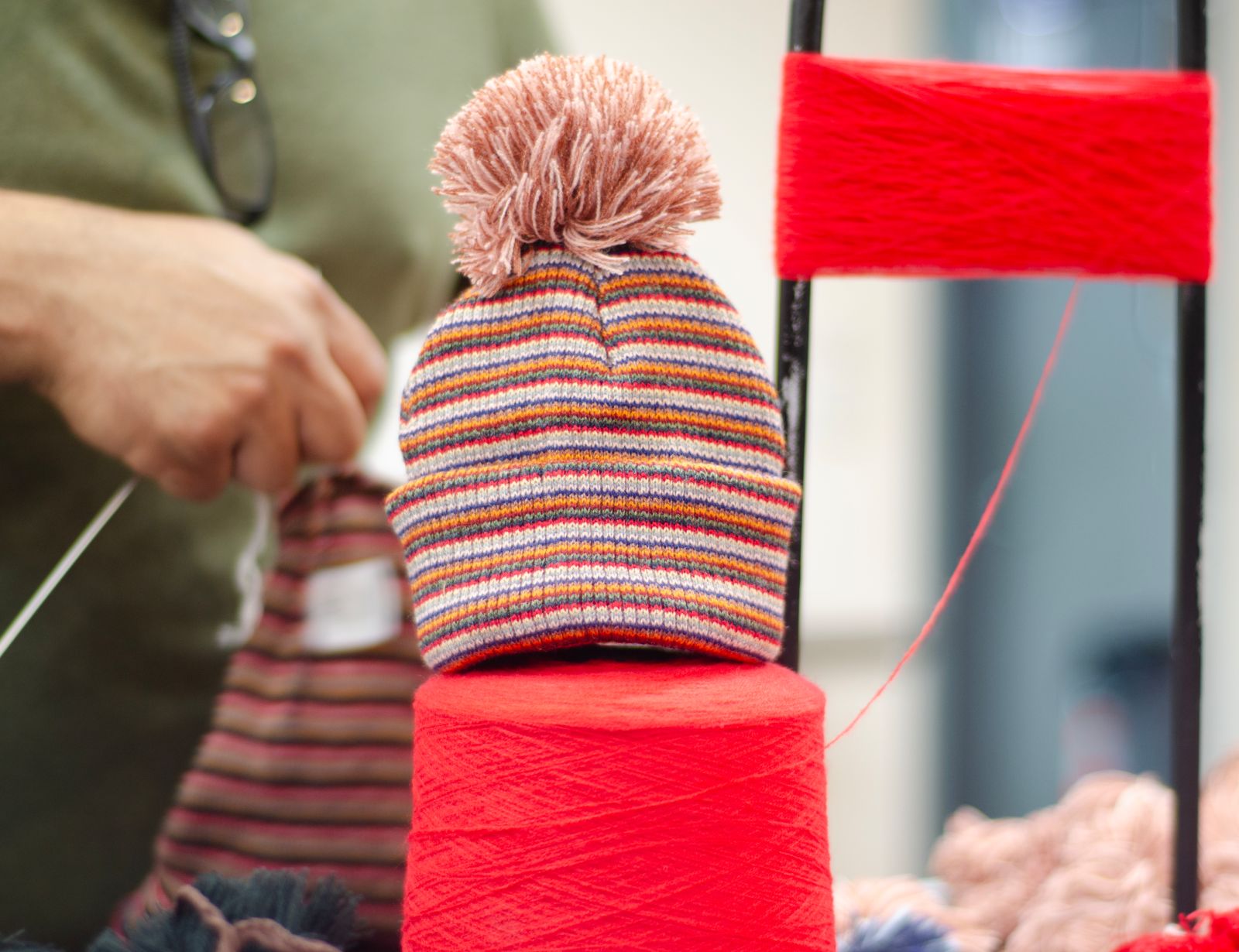 A hat with gray and orange stripes sits on a spool of brightly-colored thread. The hat has a pink pompom on top. The photo was taken in the Beanie Factory facilities in Parma, Ohio.