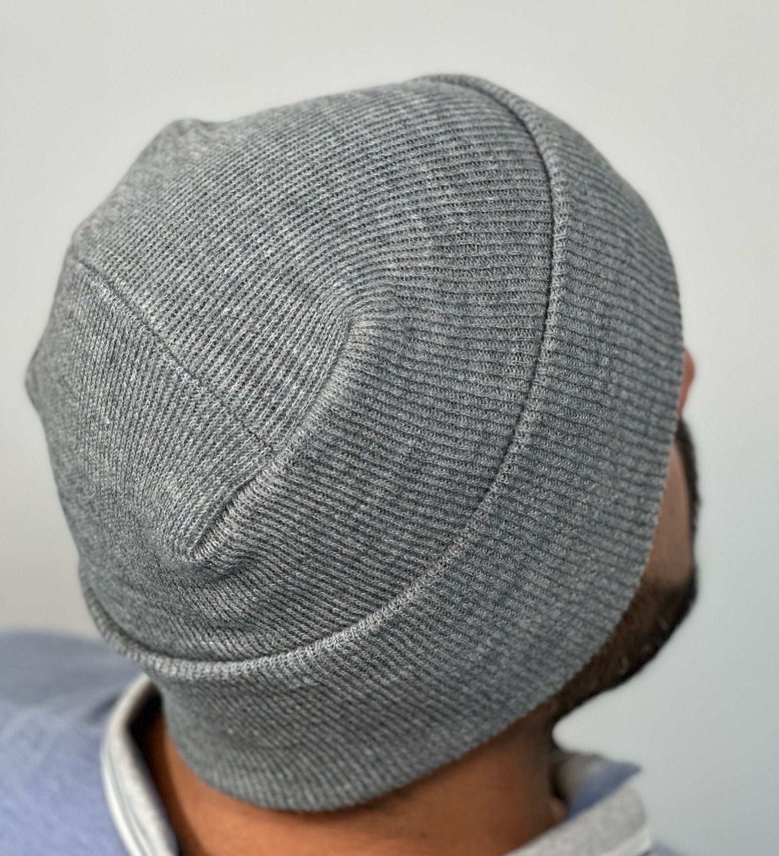 A gray hat featuring The Beanie Factory's lay-flat, irritation-free H-Stitch is modeled by a man with a black beard. The man is facing away from the camera so the H-Stitch is the focal point of the photograph.