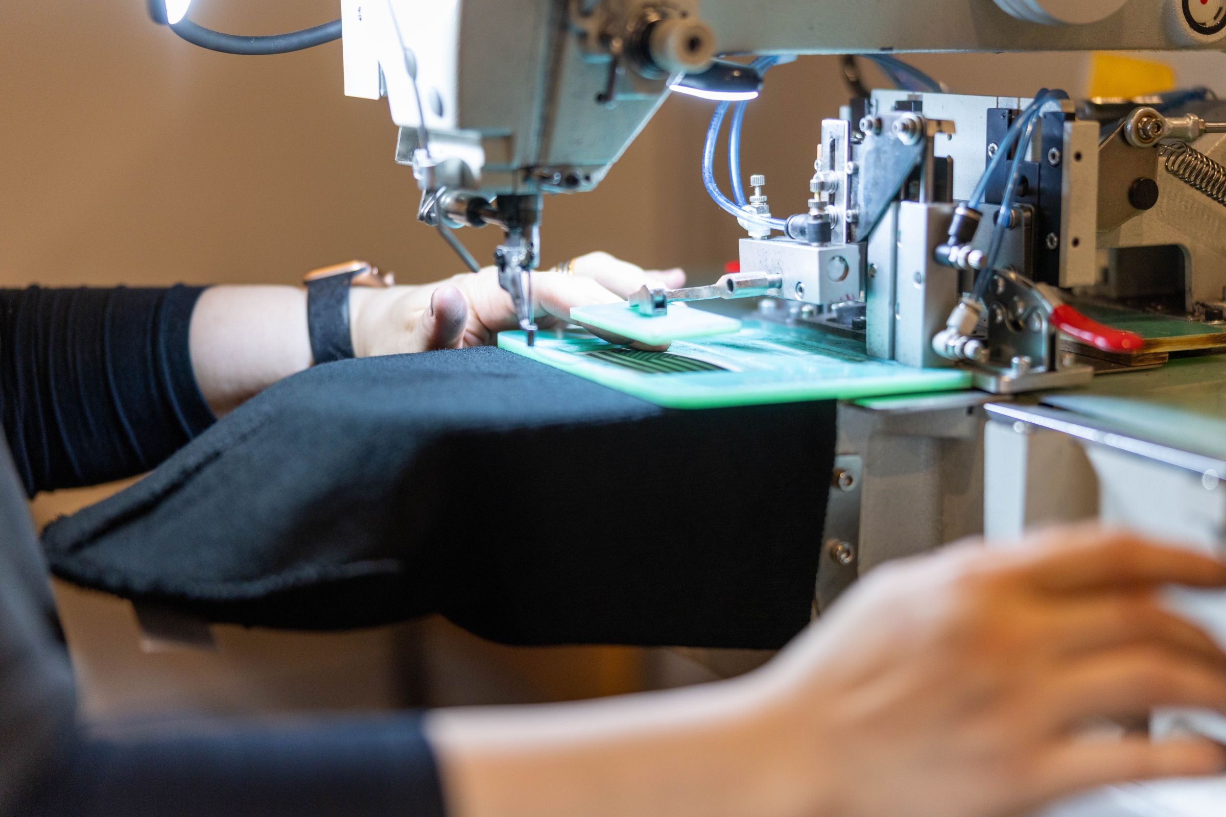 A worker at The Beanie Factory handles a black hat in a sewing machine. The image is a close-up of the sewing machine and the worker's hands.