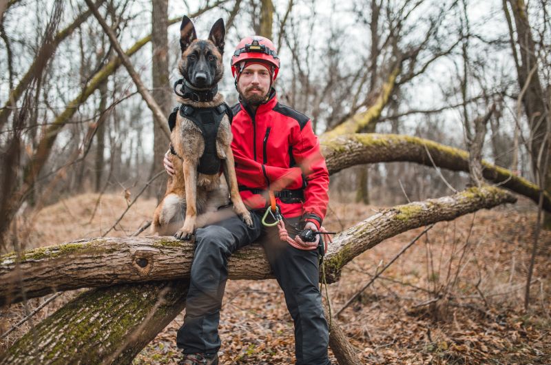 A man wearing a reflective jacket poses with a German Shepherd service dog. They are sitting on a fallen tree in the woods. The man is wearing a helmet, with The Beanie Factory's H Stitch hat underneath, showing that Beanie Pro H Stitch hats can be worn comfortably in tough conditions under helmets.