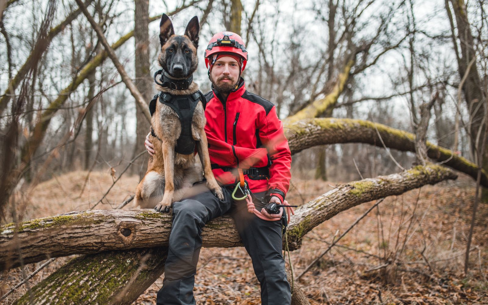 A man wearing a reflective jacket poses with a German Shepherd service dog. They are sitting on a fallen tree in the woods. The man is wearing a helmet, with The Beanie Factory's H Stitch hat underneath, showing that Beanie Pro H Stitch hats can be worn comfortably in tough conditions under helmets.