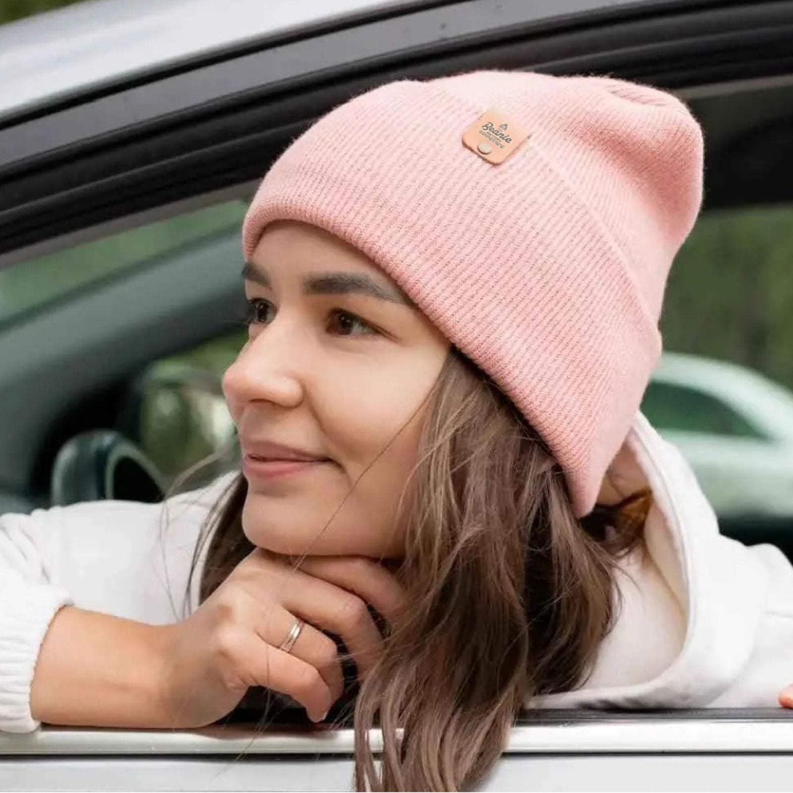 A woman looks outside a car window. She is staring off camera with a slight smile on her face, with her chin resting on her hand. She is wearing a pink Beanie Base hat from The Beanie Factory, with a custom leatherette tag on the cuff.