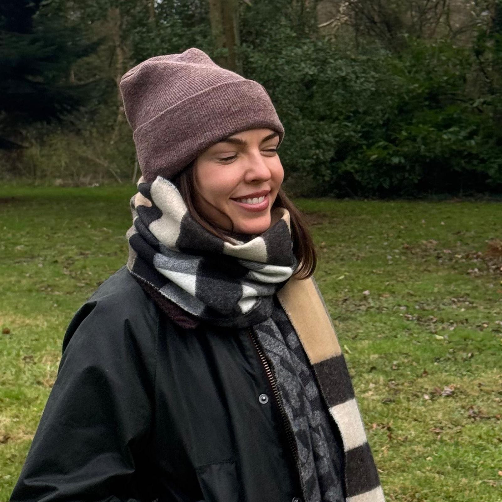 A woman smiles with her eyes closed. She is wearing a black jacket and a multi-colored scarf. She is wearing a Beanie Pro hat featuring the lay-flat, irritation-free H-Stitch.