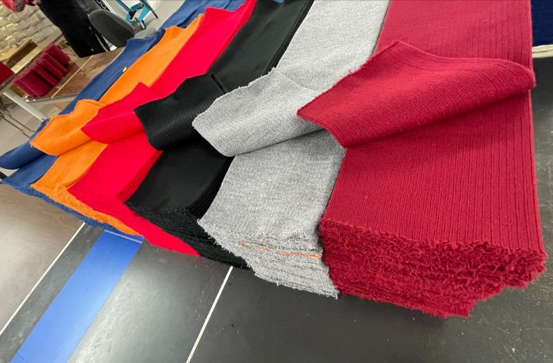 Several strips of acrylic knit fabric are on display. Red, gray, black, maroon, orange, and blue are seen. This fabric is the innovative and breathable acrylic material used to make all hats from The Beanie Factory. The innovative stitching allows for stretch without wear and without constricting elastic.