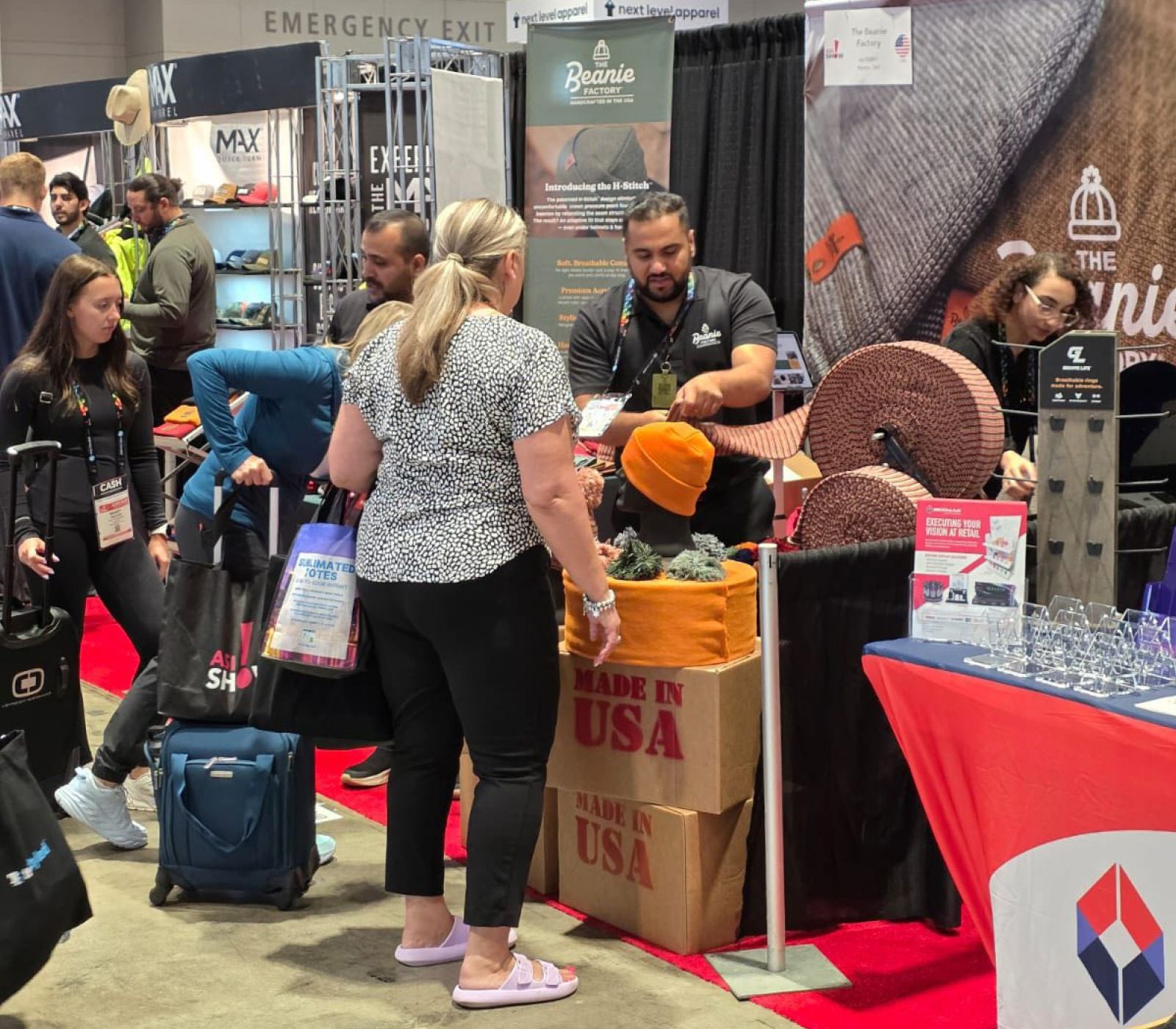 The Beanie Factory representatives at a trade show. The representative is showing a customer the quality of the fabric. There are hats and spools of fabric on display. Two boxes prominently say "Made In USA."