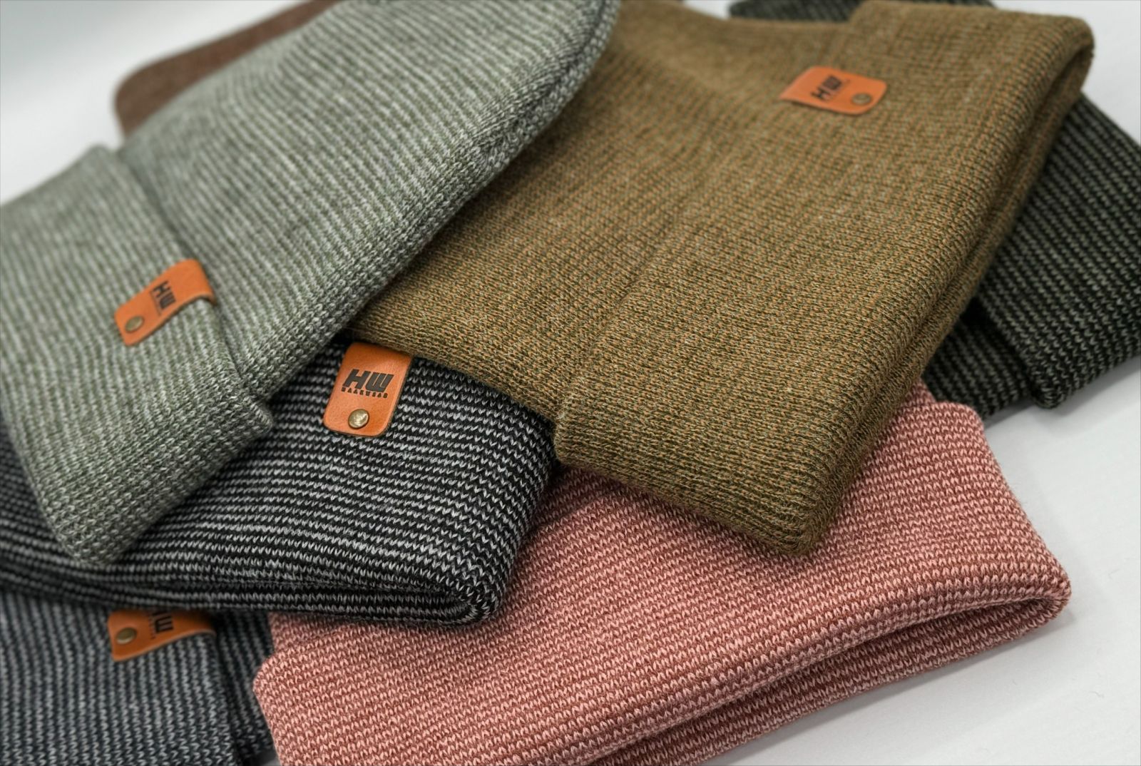 Six hats from The Beanie in different colors are piled up against a white background. The hats are customized with a branded tag that says "HW," an example of the many ways wholesale orders can be placed for customization with The Beanie Factory.